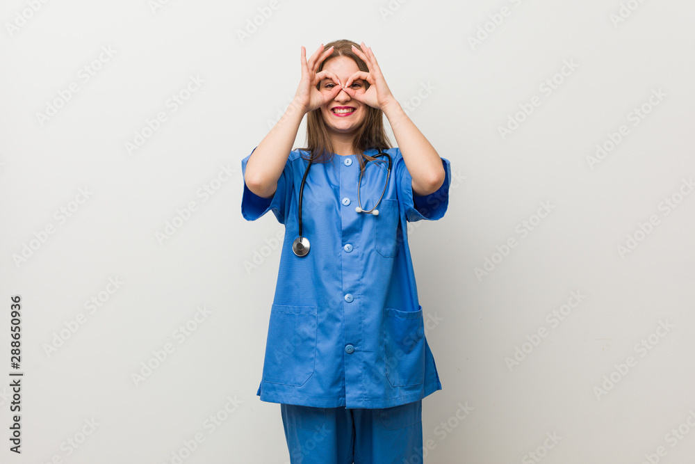 Young nurse woman against a white wall showing okay sign over eyes Young nurse woman against a white wall showing okay sign over eyes