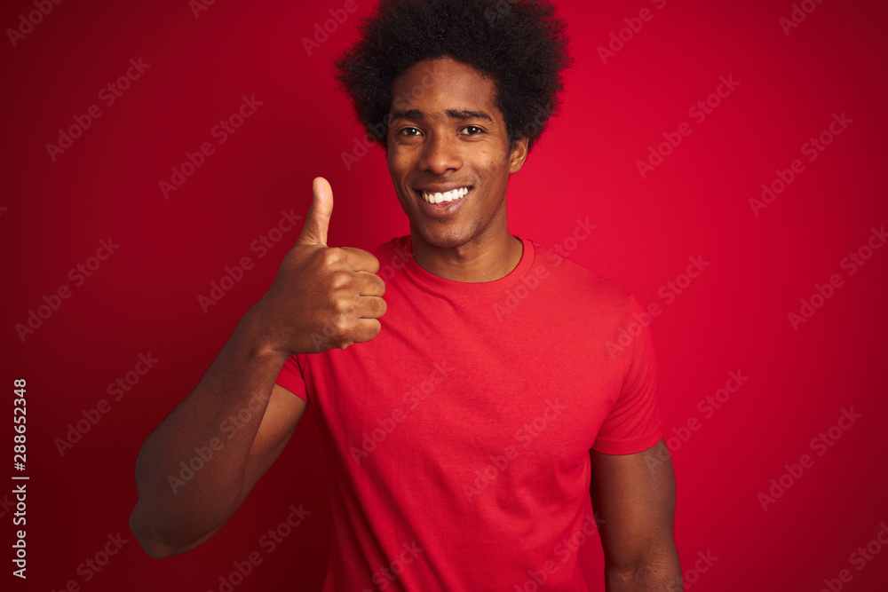 Young american man with afro hair wearing t-shirt standing over isolated red background doing happy thumbs up gesture with hand. Approving expression looking at the camera with showing success. Young american man with afro hair wearing t-shirt standing over isolated red background doing happy thumbs up gesture with hand. Approving expression looking at the camera with showing success.