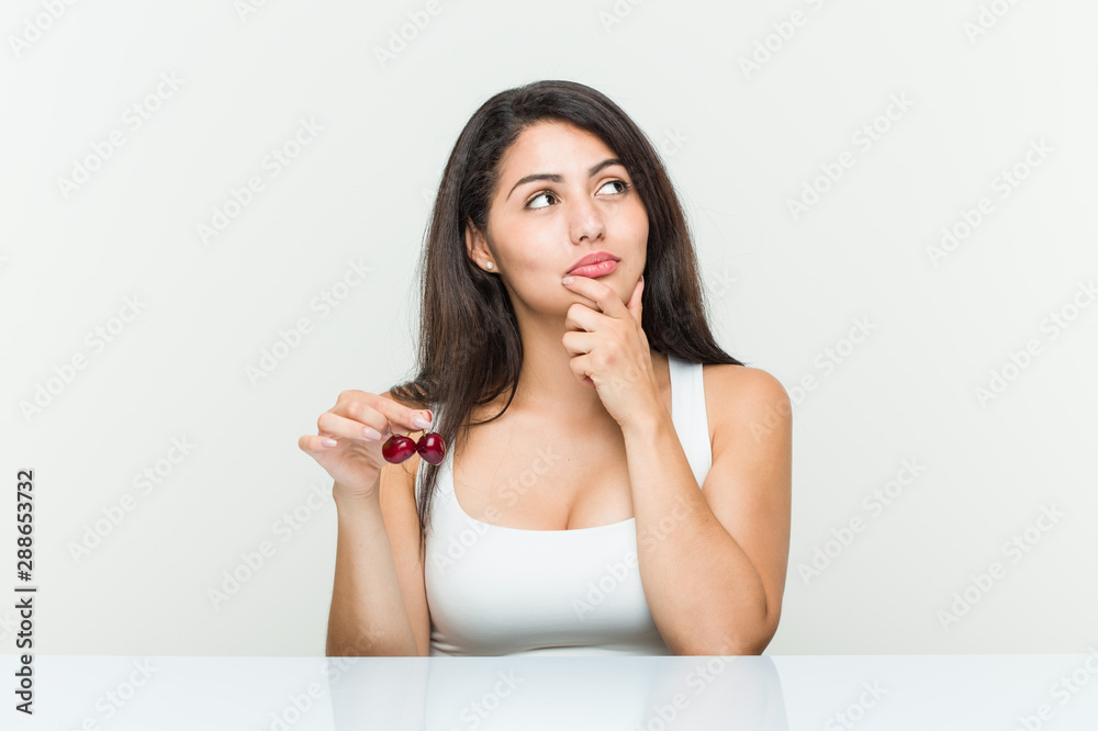 Young hispanic woman holding cherries looking sideways with doubtful and skeptical expression. Young hispanic woman holding cherries looking sideways with doubtful and skeptical expression.