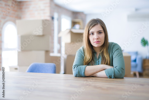 Free Download Young woman sitting on the table with cardboard boxes behind her moving to new home skeptic and nervous, disapproving expression on face with crossed arms. Negative person. - GFXLoad