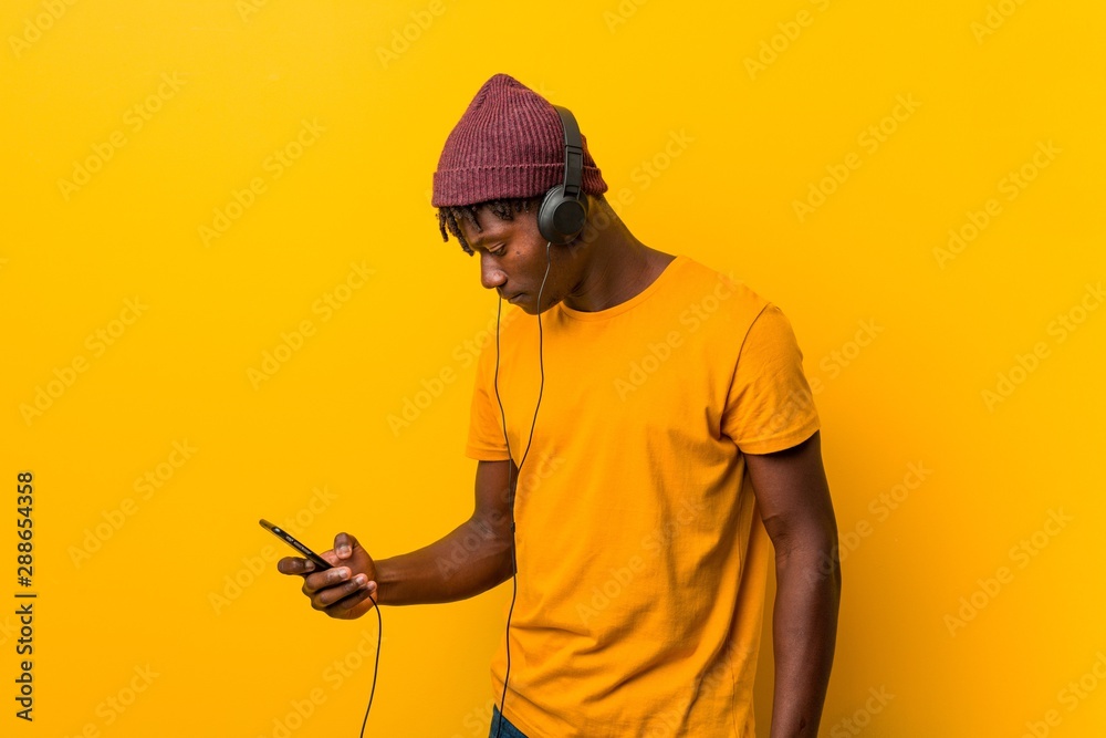 Young african man standing against a yellow background wearing a hat listening to music with a phone Young african man standing against a yellow background wearing a hat listening to music with a phone