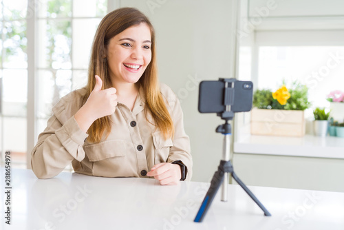 Beautiful young woman doing online video call using smartphone webcam doing happy thumbs up gesture with hand. Approving expression looking at the camera with showing success. photo