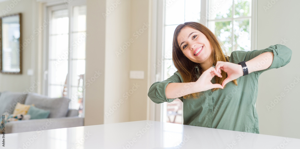 Beautiful young woman at home smiling in love showing heart symbol and shape with hands. Romantic concept.