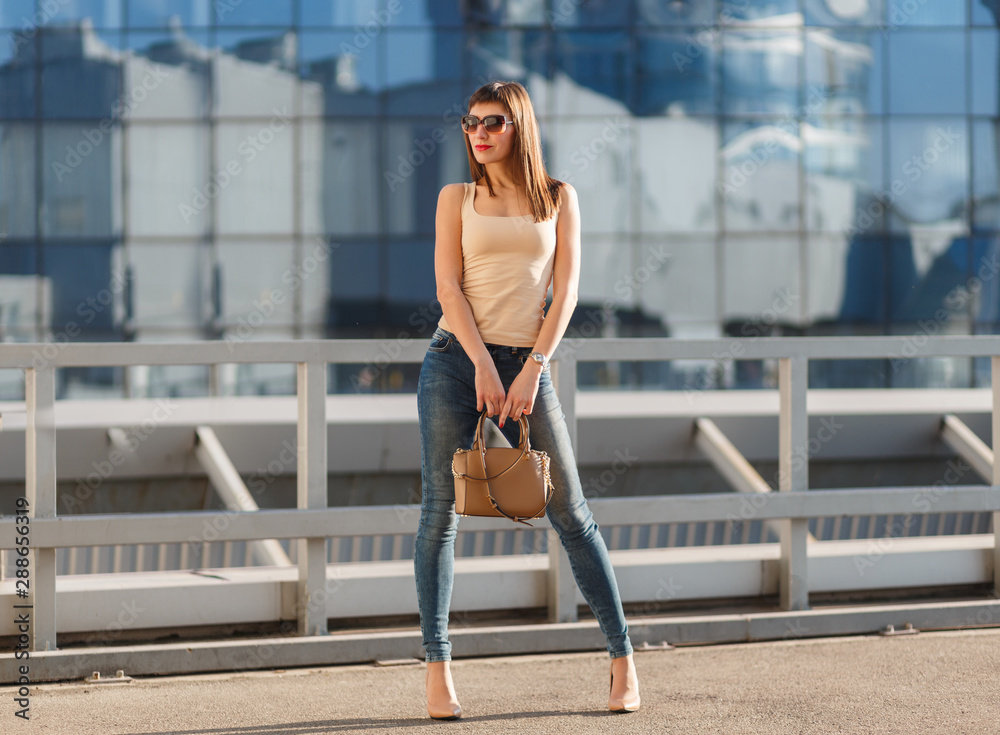 Portrait of beautiful brunette young woman in nice beige shirt, denim jeans  and sunglasses. Genuine leather bag, high heels shoes. Spring autumn  fashion trendy photo of lady on urban background. foto de