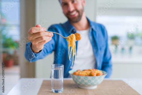 Handsome man eating pasta with meatballs and tomato sauce at home while smiling at the camera