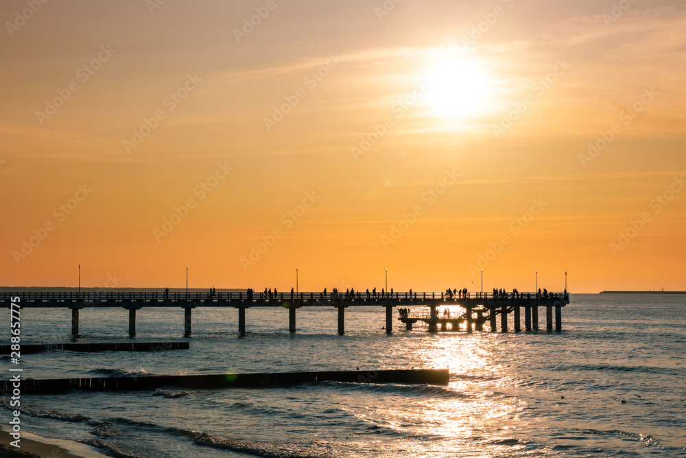silhouettes of people walking on a pier by the sea against a sunset orange sky