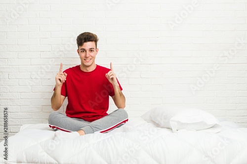 Young teenager student man on the bed indicates with both fore fingers up showing a blank space.