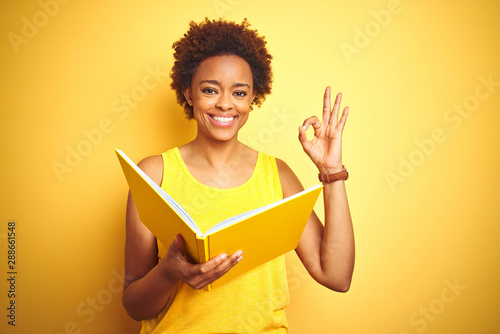 African american woman reading a book over yellow isolated background doing ok sign with fingers, excellent symbol