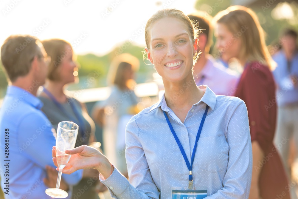 Portrait of young smiling businesswoman standing while holding wine glass against colleagues at success party on rooftop Portrait of young smiling businesswoman standing while holding wine glass against colleagues at success party on rooftop