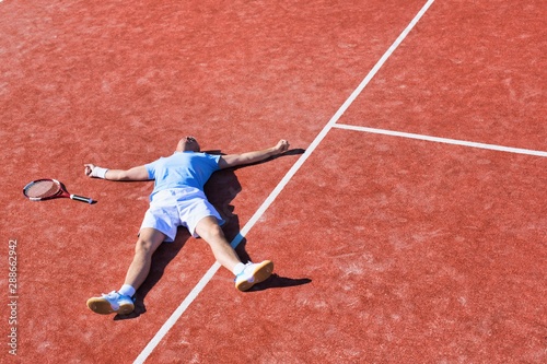 Full length of disappointed mature man lying by tennis racket on court during summer