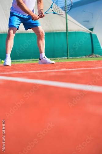 Confident mature man hitting tennis ball with racket on red court