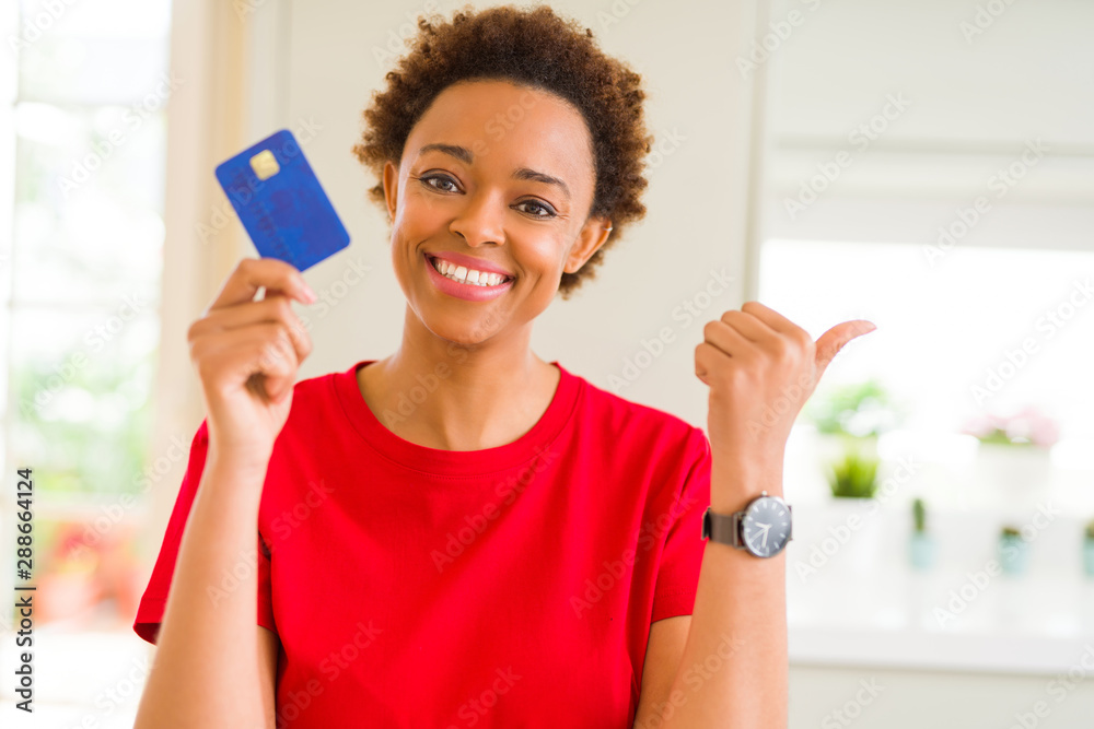 Young african american woman holding credit card pointing and showing with thumb up to the side with happy face smiling Young african american woman holding credit card pointing and showing with thumb up to the side with happy face smiling