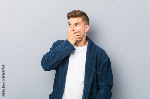 Young caucasian man wearing pajama thoughtful looking to a copy space covering mouth with hand.