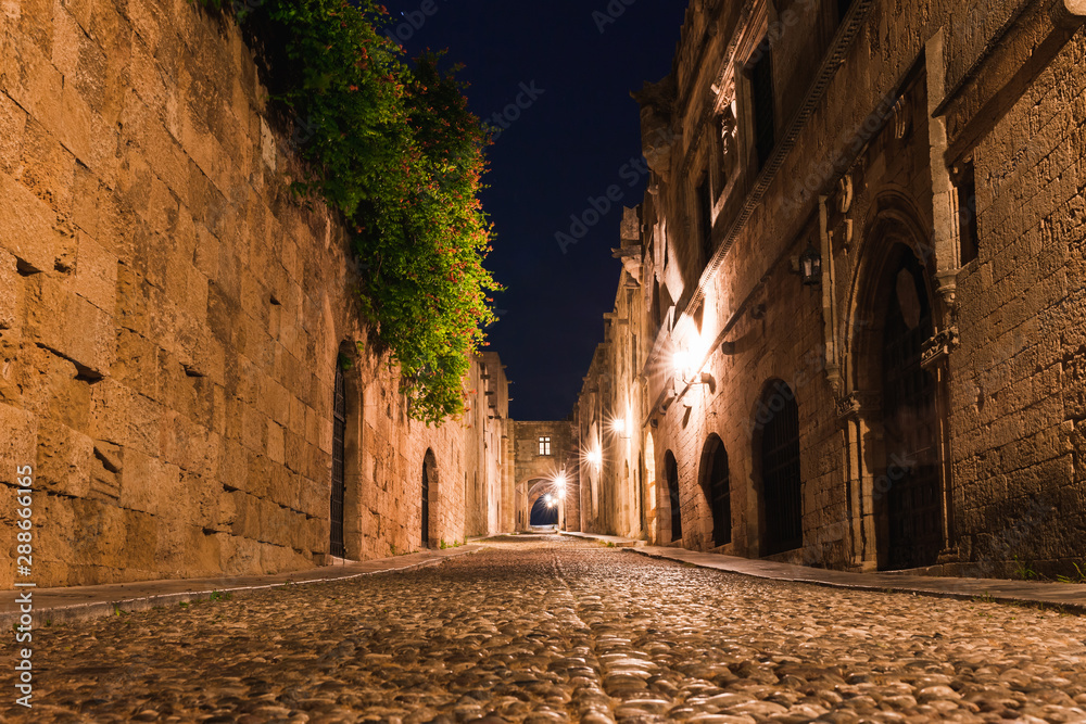 Night photo of ancient street of the Knights in Rhodes city on Rhodes island, Dodecanese, Greece. Stone walls and bright night lights. Famous tourist destination in South Europe