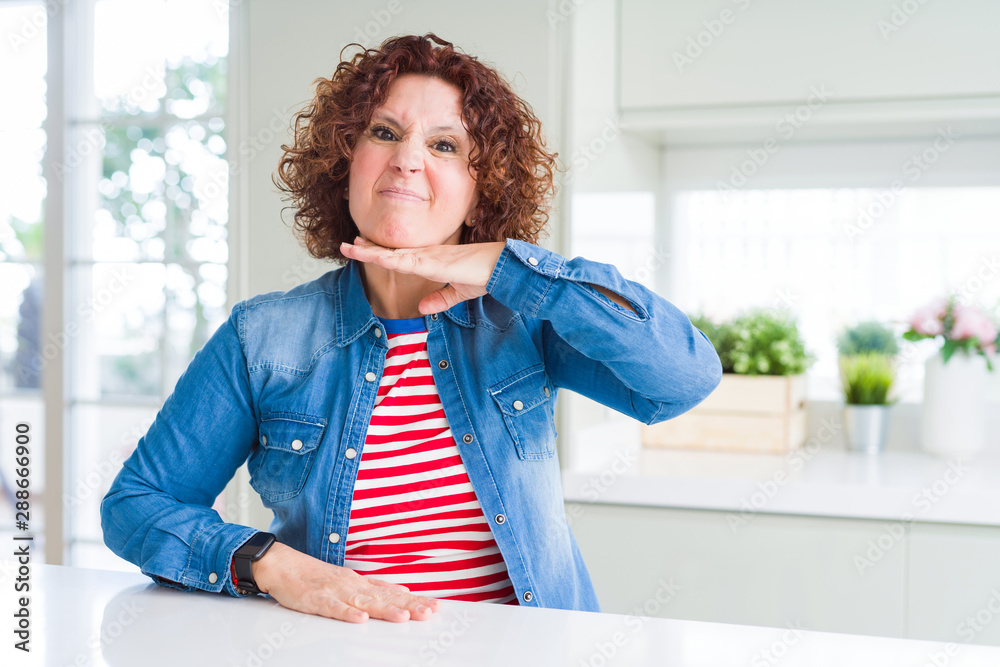 Middle age senior woman with curly hair wearing denim jacket at home cutting throat with hand as knife, threaten aggression with furious violence