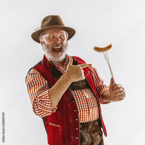 Portrait of Oktoberfest senior man in hat, wearing the traditional Bavarian clothes. Male full-length shot at studio on white background. The celebration, holidays, festival concept. Eating sausage.