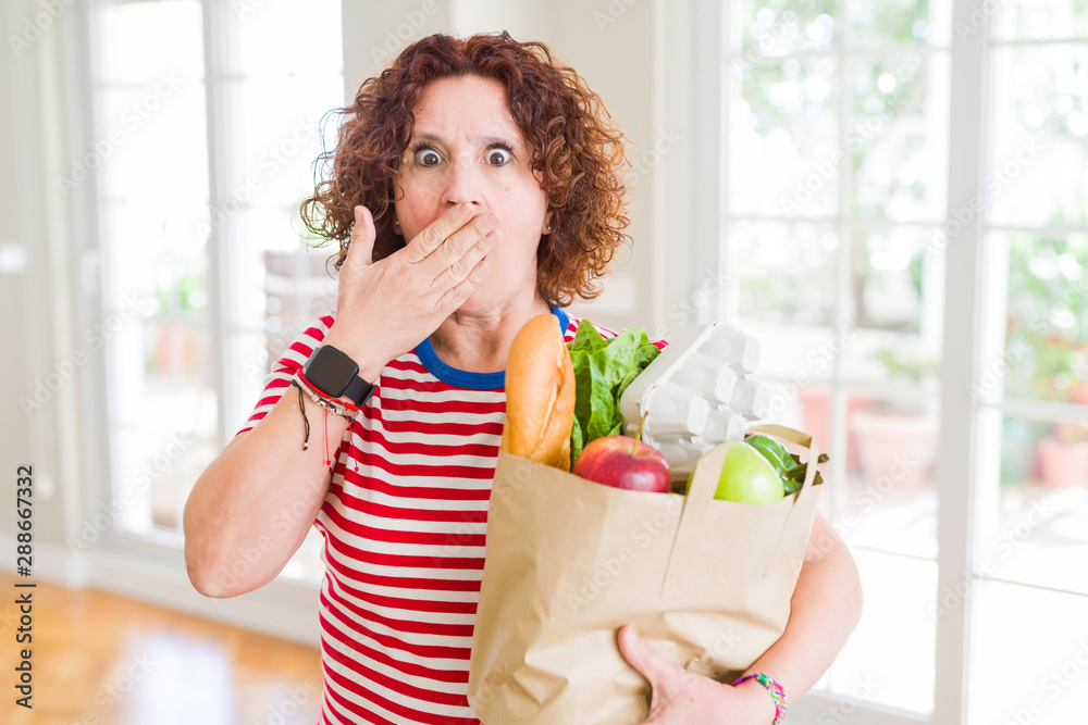 Senior woman holding paper bag full of fresh groceries from the supermarket cover mouth with hand shocked with shame for mistake, expression of fear, scared in silence, secret concept Senior woman holding paper bag full of fresh groceries from the supermarket cover mouth with hand shocked with shame for mistake, expression of fear, scared in silence, secret concept