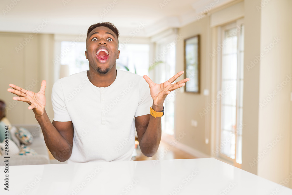 Handsome african american man on white table at home celebrating crazy and amazed for success with arms raised and open eyes screaming excited. Winner concept Handsome african american man on white table at home celebrating crazy and amazed for success with arms raised and open eyes screaming excited. Winner concept