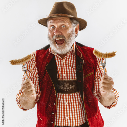Portrait of Oktoberfest senior man in hat, wearing the traditional Bavarian clothes. Male full-length shot at studio on white background. The celebration, holidays, festival concept. Eating sausages.
