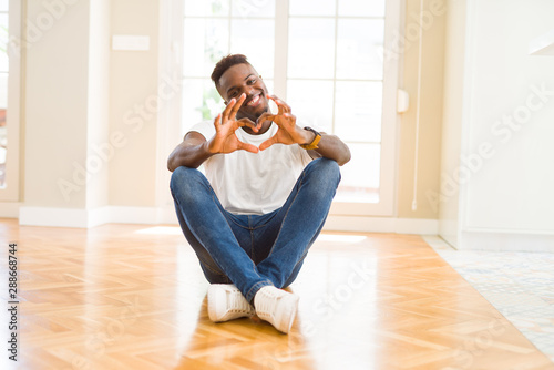 Handsome african american man sitting on the floor at home smiling in love showing heart symbol and shape with hands. Romantic concept.