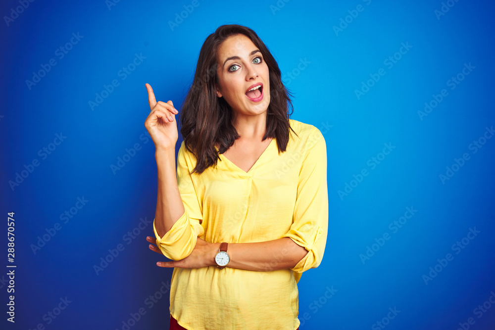 Young beautiful woman wearing yellow t-shirt standing over blue isolated background with a big smile on face, pointing with hand and finger to the side looking at the camera.