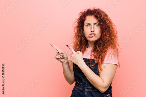 Young pretty ginger redhead woman wearing a jeans dungaree shocked pointing with index fingers to a copy space.