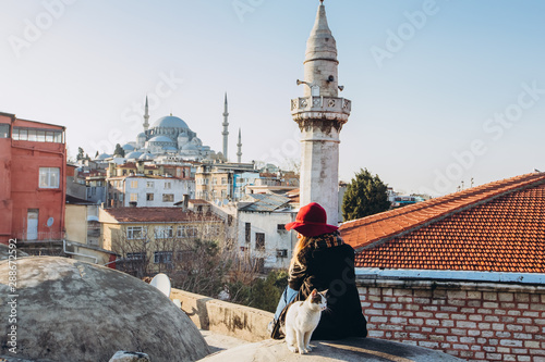 Blond woman stroking a cat on the roof with a view to the mosque, Istanbul, Turkey. Girl in a hat sits on the roof in Istanbul, sunny autumn day. Traveler girl walks through winter Istanbul.