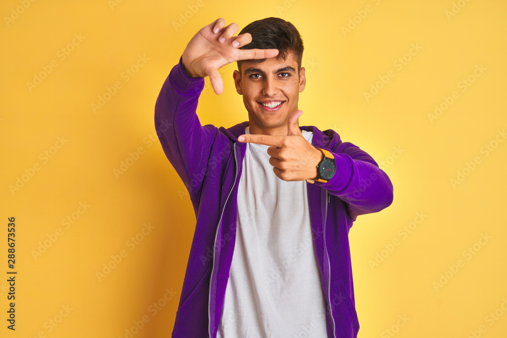 Young indian man wearing purple sweatshirt standing over isolated yellow background smiling making frame with hands and fingers with happy face. Creativity and photography concept. Young indian man wearing purple sweatshirt standing over isolated yellow background smiling making frame with hands and fingers with happy face. Creativity and photography concept.