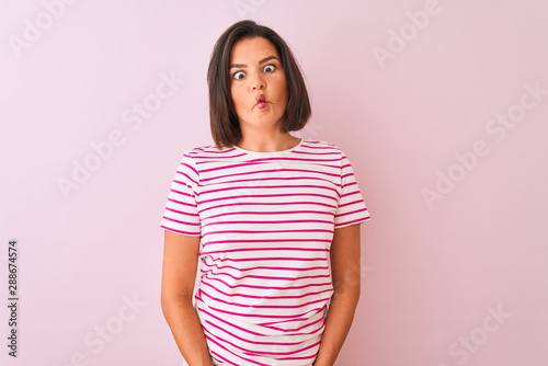 Young beautiful woman wearing striped t-shirt standing over isolated pink background making fish face with lips, crazy and comical gesture. Funny expression.