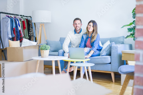 Young beautiful couple sitting on the sofa drinking cup of coffee using laptop at new home around cardboard boxes