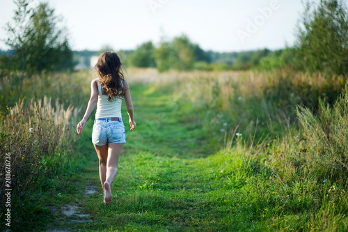 girl in denim shorts with her hair running away barefoot along a country road. copy space © Вячеслав Думчев