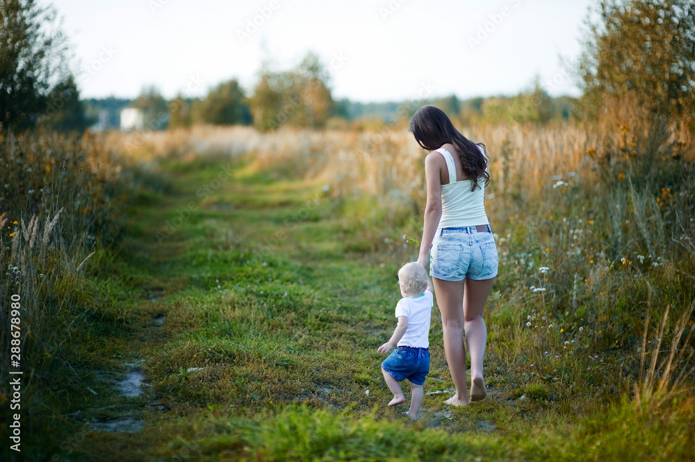 young mother walks with her little daughter barefoot on a country road at sunset, view from the back. The nanny walks with the child. copy space. toned young mother walks with her little daughter barefoot on a country road at sunset, view from the back. The nanny walks with the child. copy space. toned
