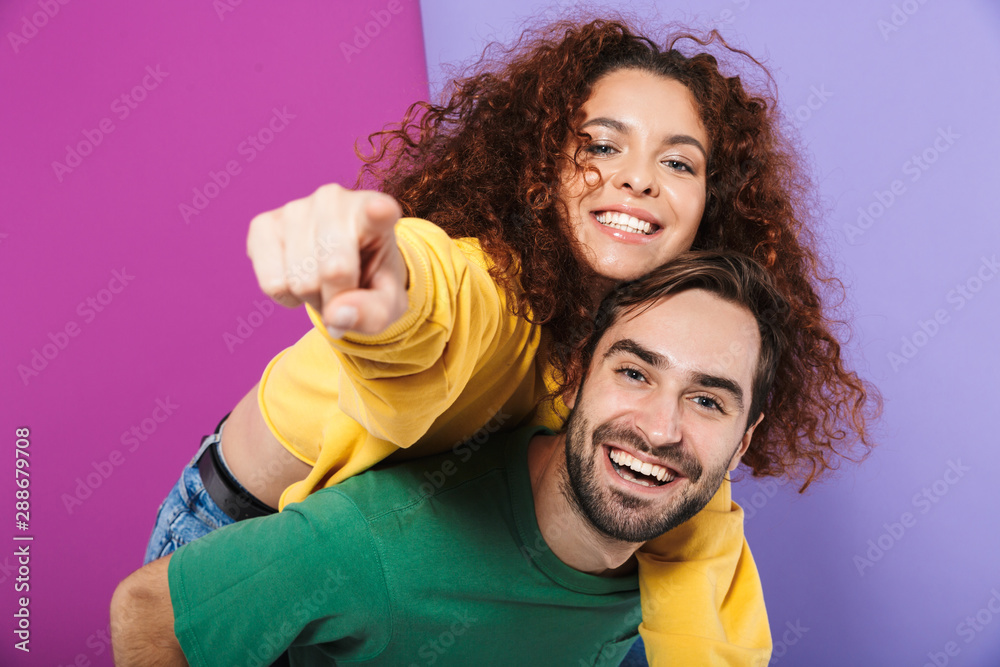 Fototapeta premium Portrait of excited smiling couple man and woman pointing finger at camera while doing piggyback ride