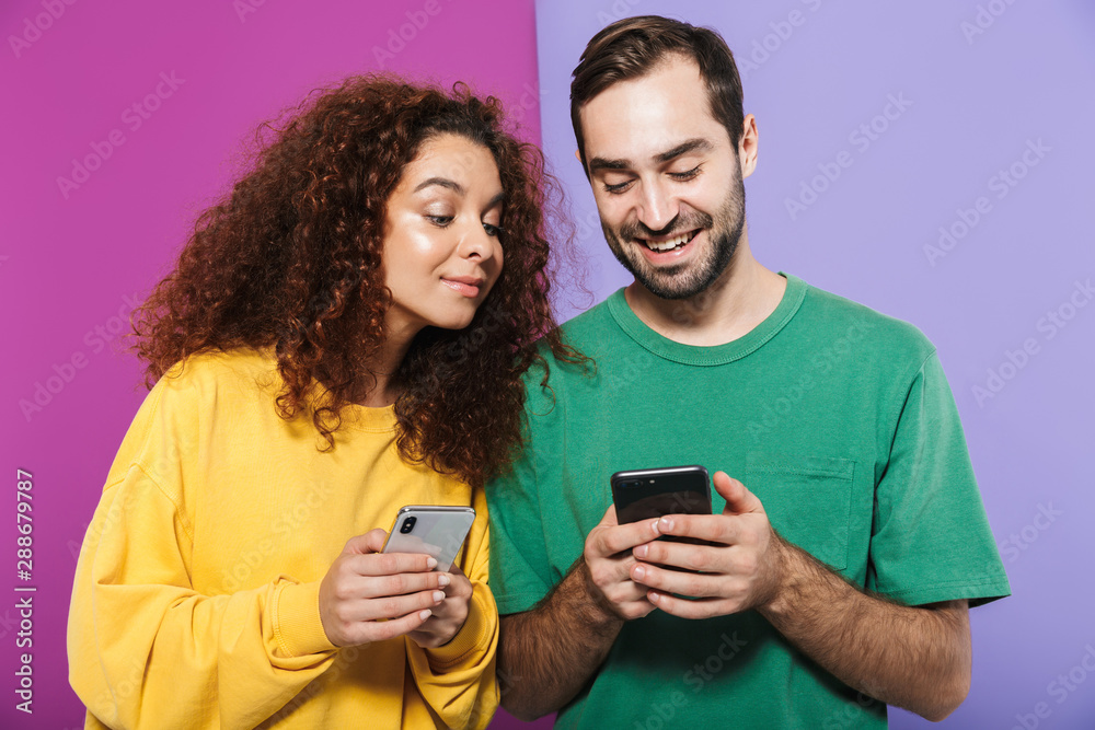 Fototapeta premium Portrait of joyful caucasian couple man and woman in colorful clothing smiling and holding cellphones together
