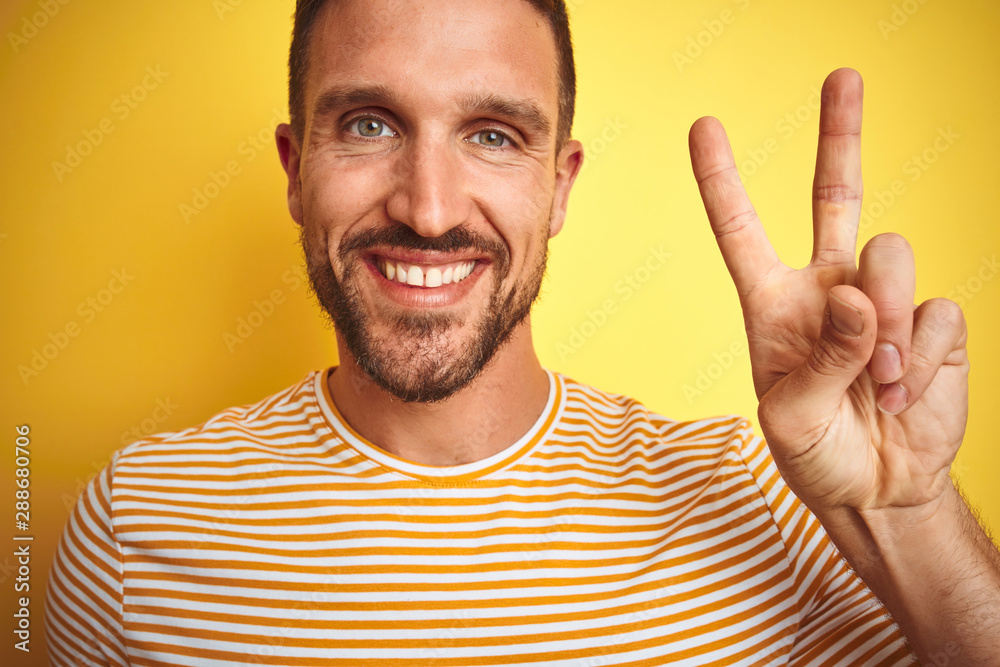 Close up of young handsome man wearing casual t-shirt over yellow isolated background smiling with happy face winking at the camera doing victory sign. Number two. Close up of young handsome man wearing casual t-shirt over yellow isolated background smiling with happy face winking at the camera doing victory sign. Number two.