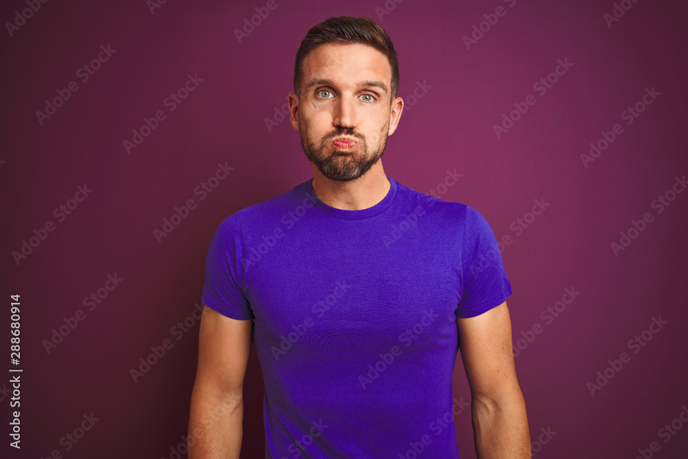 Young man wearing casual purple t-shirt over lilac isolated background puffing cheeks with funny face. Mouth inflated with air, crazy expression. Young man wearing casual purple t-shirt over lilac isolated background puffing cheeks with funny face. Mouth inflated with air, crazy expression.