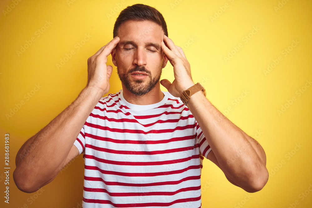 Young handsome man wearing casual red striped t-shirt over yellow isolated background with hand on head for pain in head because stress. Suffering migraine. Young handsome man wearing casual red striped t-shirt over yellow isolated background with hand on head for pain in head because stress. Suffering migraine.