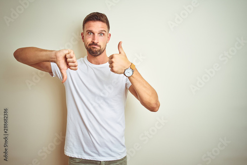 Bilde på lerret Young handsome man wearing casual white t-shirt over isolated background Doing thumbs up and down, disagreement and agreement expression