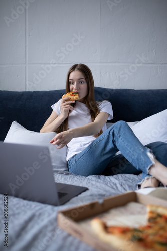 Happy young woman eating slice of hot pizza at home and watching film on laptop