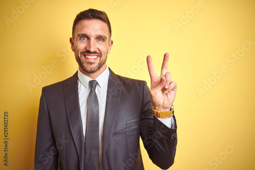 Young handsome business man over yellow isolated background smiling with happy face winking at the camera doing victory sign. Number two.