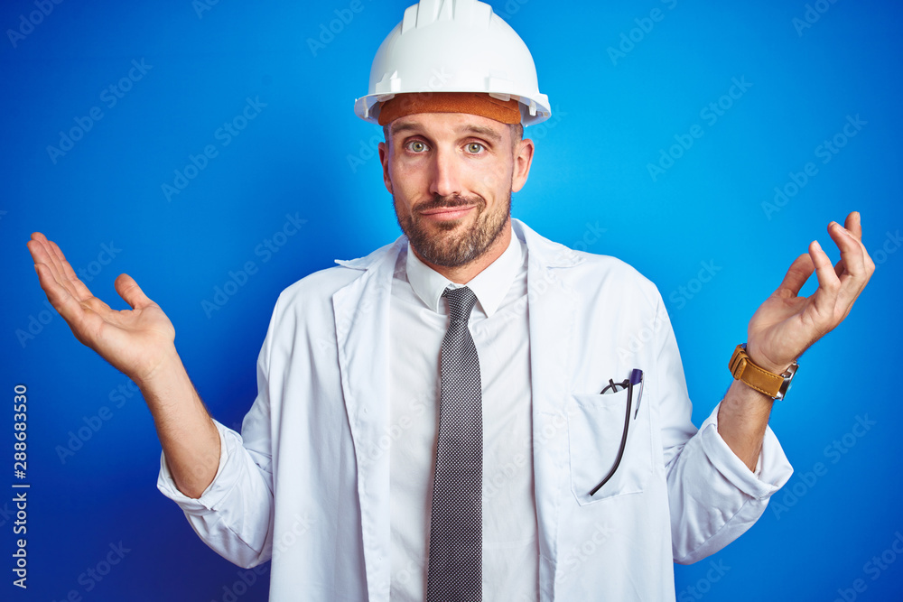 Young handsome engineer man wearing safety helmet over blue isolated background clueless and confused expression with arms and hands raised. Doubt concept.