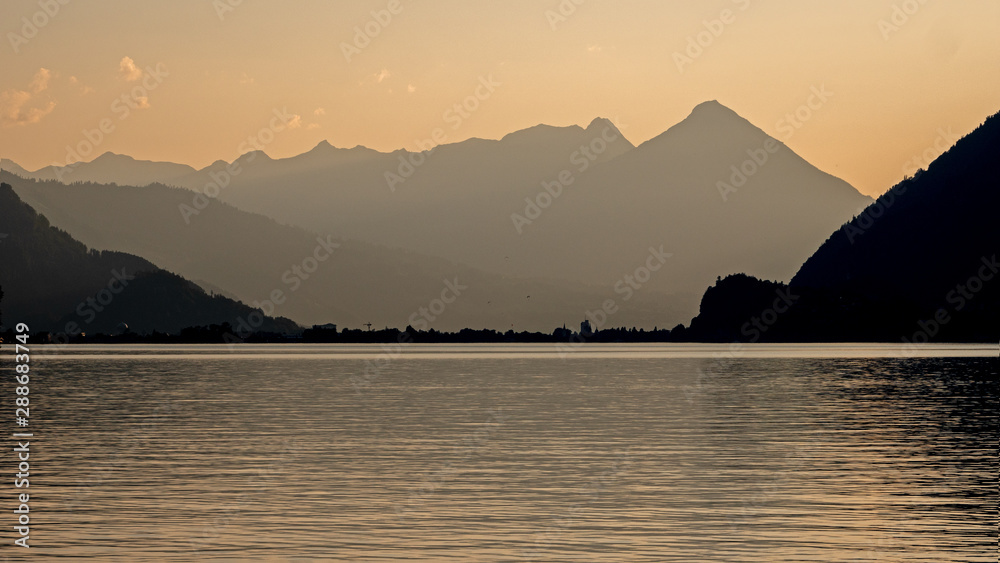 Sunset looking down the lake of Brienz in Switzerland. The high mountains cast a silhouette against the orange sky and reflections in the water of the alpine lake. 