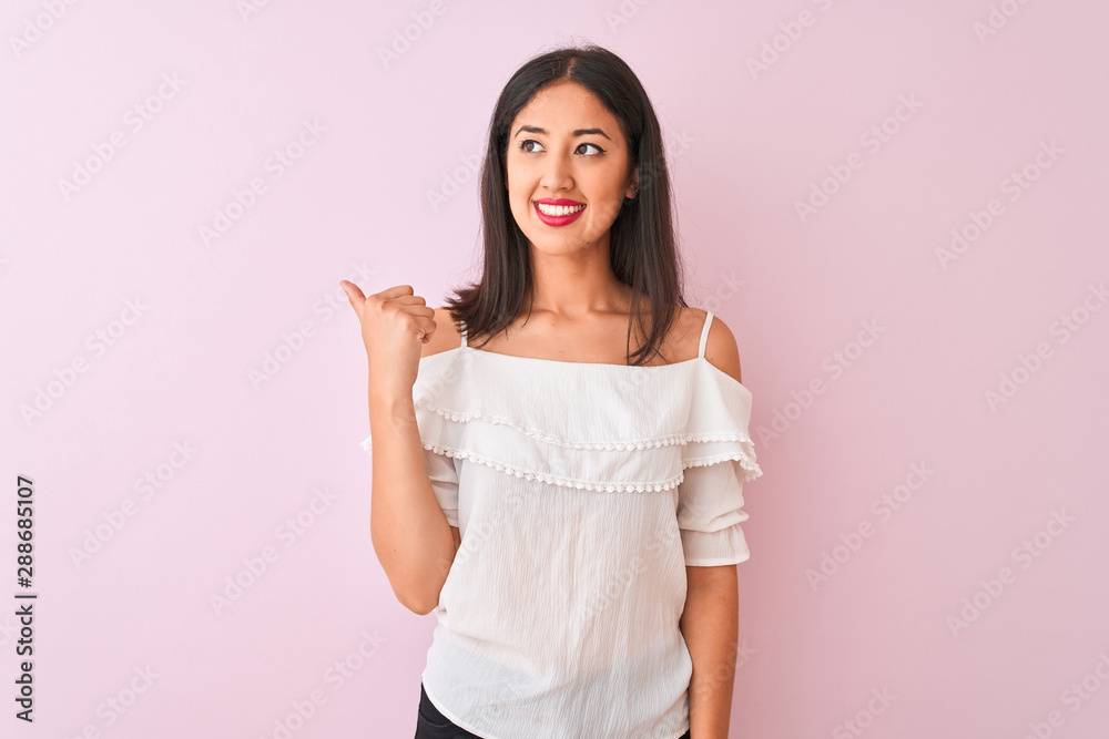 Beautiful chinese woman wearing white t-shirt standing over isolated pink background smiling with happy face looking and pointing to the side with thumb up. Beautiful chinese woman wearing white t-shirt standing over isolated pink background smiling with happy face looking and pointing to the side with thumb up.