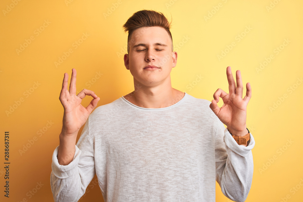 Young handsome man wearing white casual t-shirt standing over isolated yellow background relax and smiling with eyes closed doing meditation gesture with fingers. Yoga concept. Young handsome man wearing white casual t-shirt standing over isolated yellow background relax and smiling with eyes closed doing meditation gesture with fingers. Yoga concept.