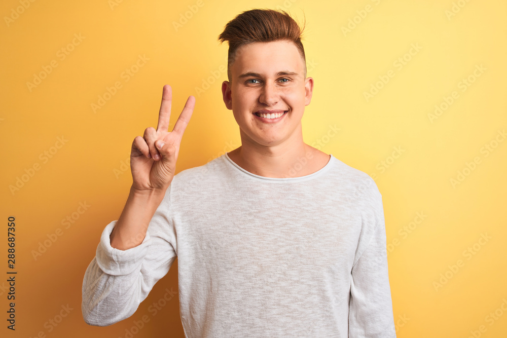 Young handsome man wearing white casual t-shirt standing over isolated yellow background showing and pointing up with fingers number two while smiling confident and happy. Young handsome man wearing white casual t-shirt standing over isolated yellow background showing and pointing up with fingers number two while smiling confident and happy.