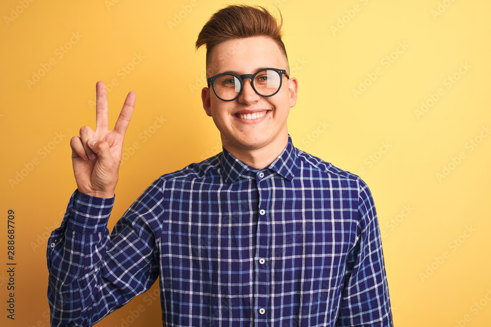 Young handsome man wearing casual shirt and glasses over isolated yellow background smiling with happy face winking at the camera doing victory sign. Number two.