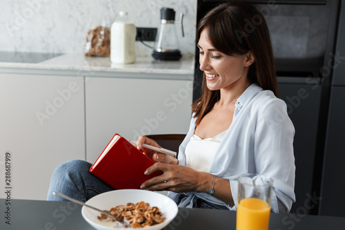 Canvastavla Happy cheery woman indoors at home at the kitchen have a breakfast eat corn flakes while writing notes in notebook