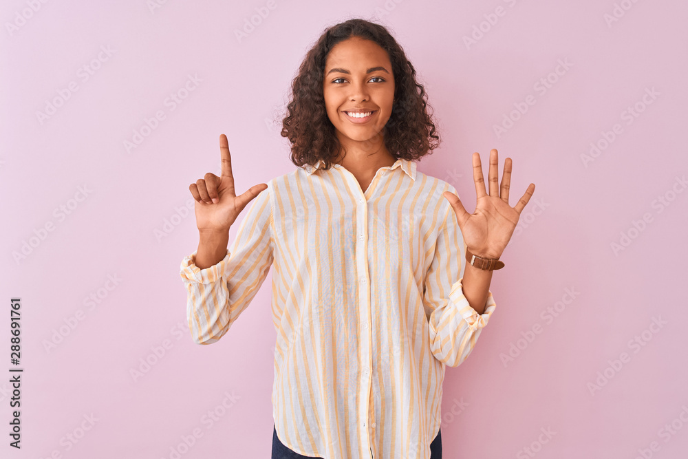 Young brazilian woman wearing striped shirt standing over isolated pink background showing and pointing up with fingers number seven while smiling confident and happy. Young brazilian woman wearing striped shirt standing over isolated pink background showing and pointing up with fingers number seven while smiling confident and happy.