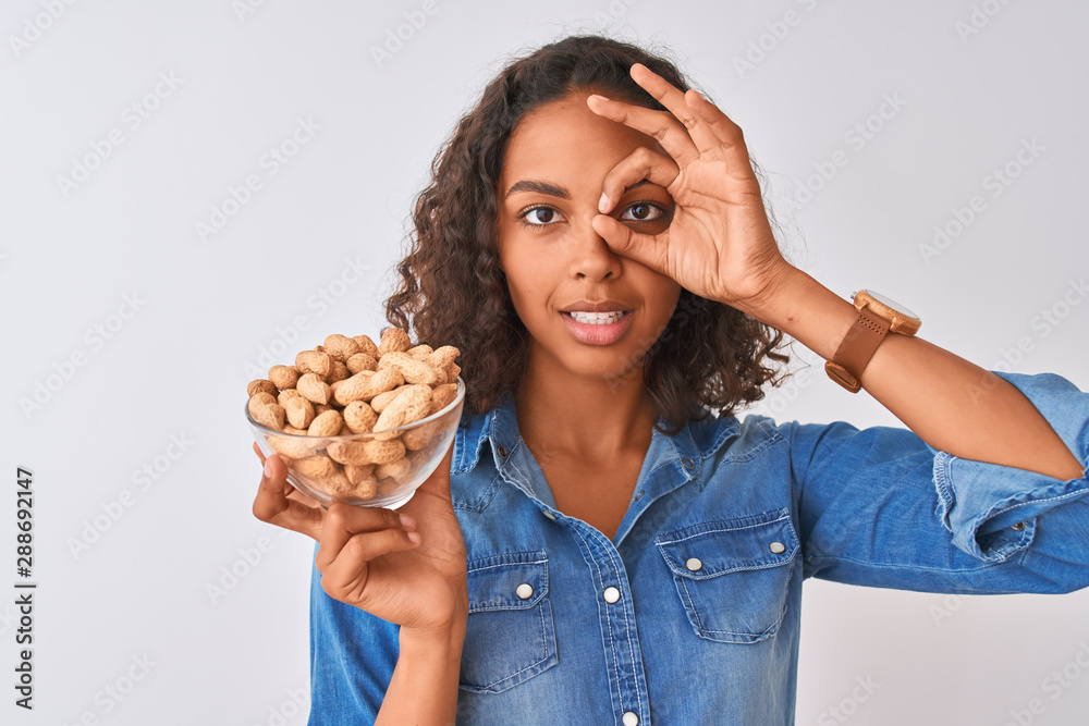 Young brazilian woman holding bowl with peanuts standing over isolated white background with happy face smiling doing ok sign with hand on eye looking through fingers Young brazilian woman holding bowl with peanuts standing over isolated white background with happy face smiling doing ok sign with hand on eye looking through fingers