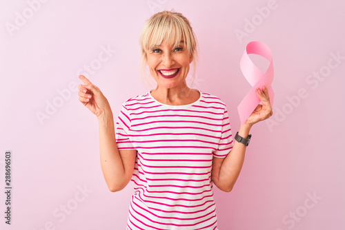 Middle age woman holding cancer ribbon standing over isolated pink background very happy pointing with hand and finger to the side